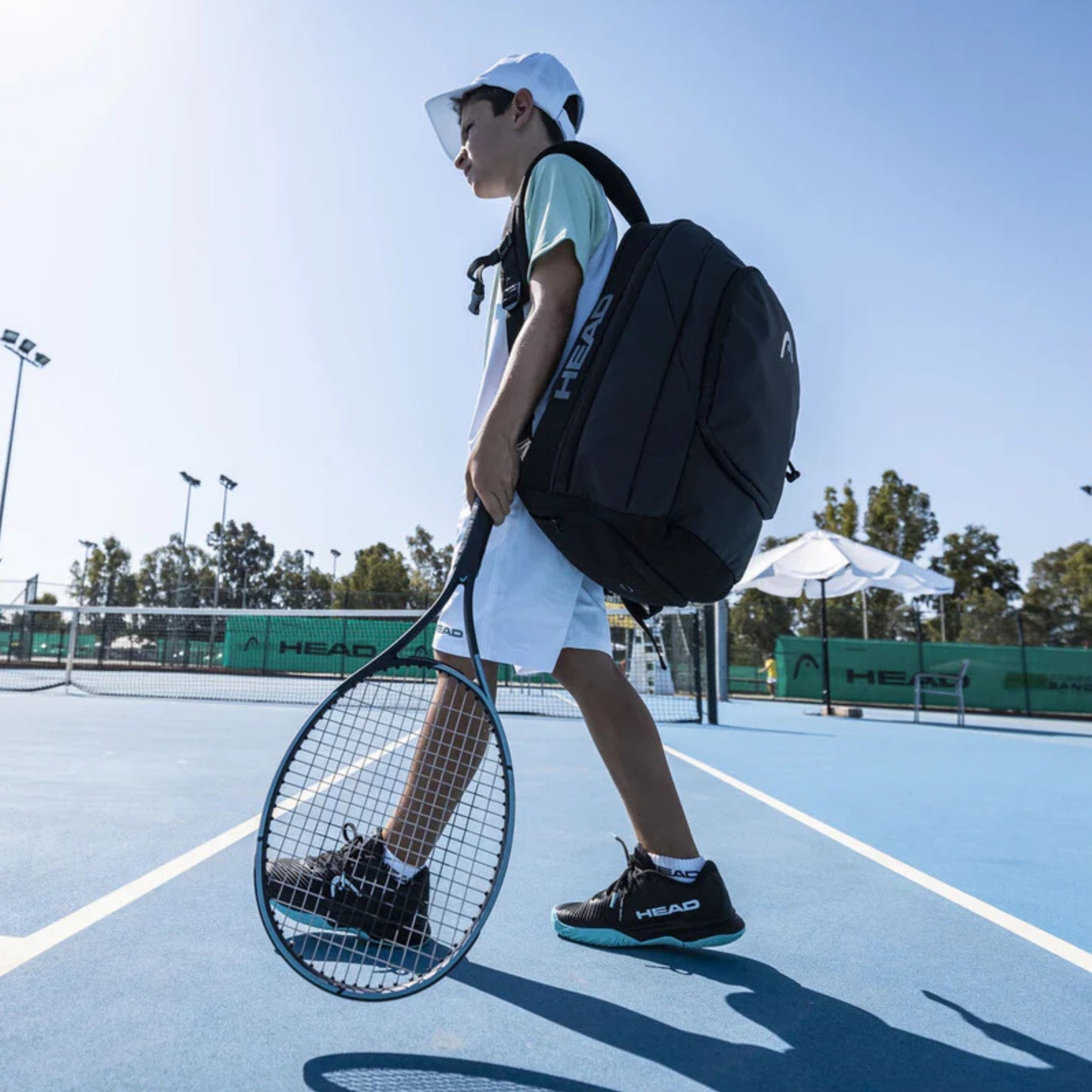 Person holding a blue and black Head tennis racket on a tennis court with a backpack, wearing a white cap and shirt.