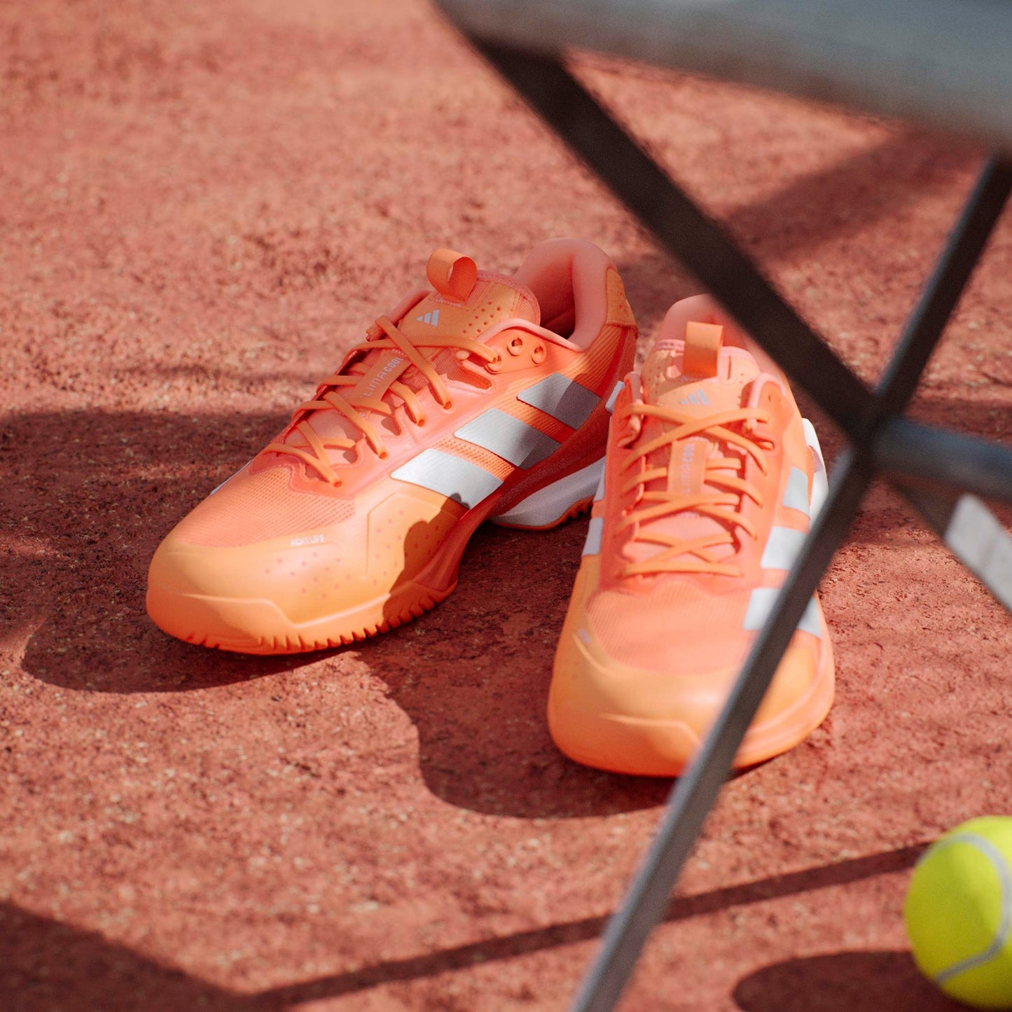 Orange tennis shoes on a red clay court with a tennis racket and ball.