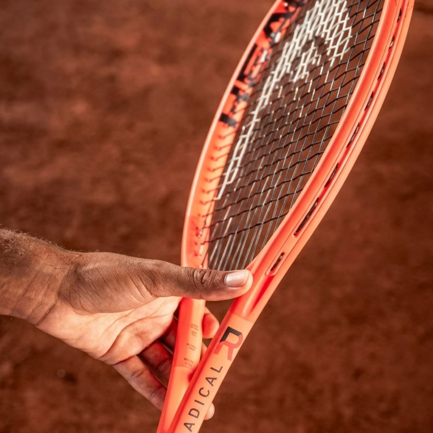 A close-up of a Head tennis racket with a bright orange frame and black-and-white string bed, held by a person's hand against a blurred brown clay court background.