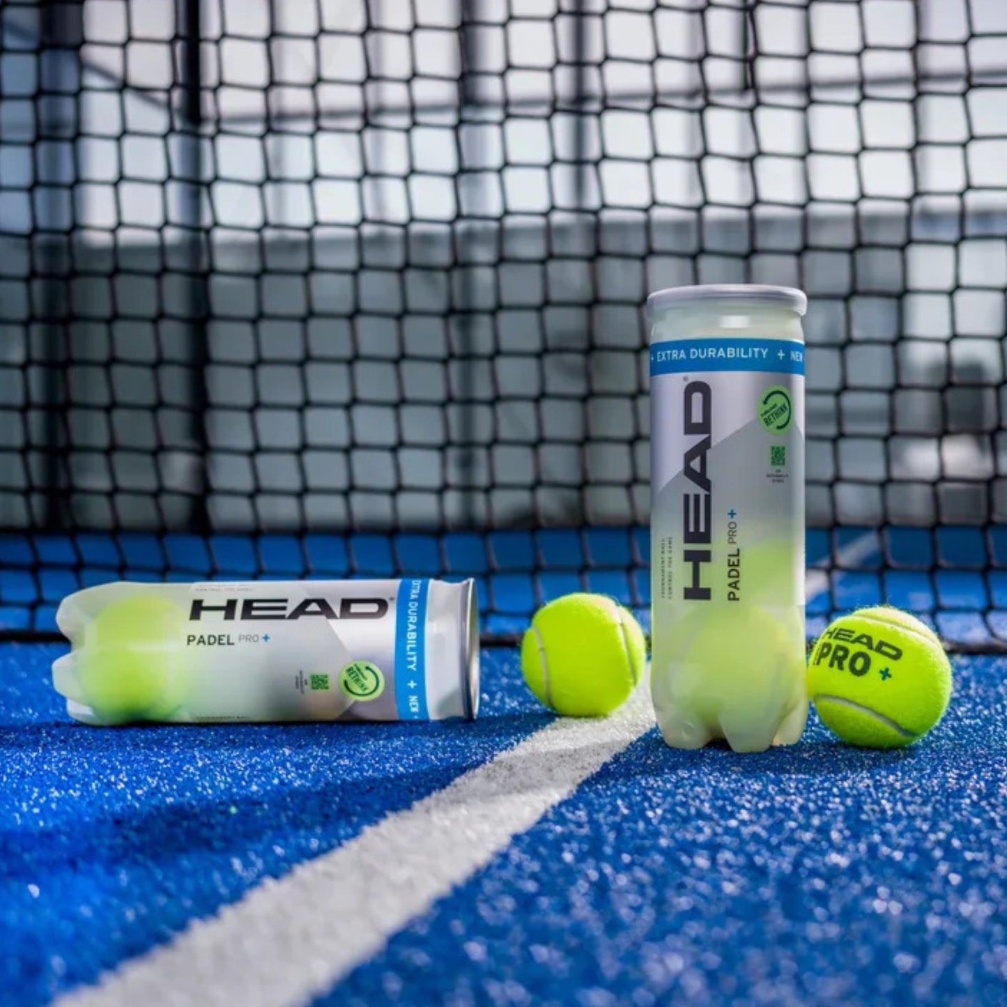 Two cans of HEAD tennis balls on a tennis court with a net in the background.