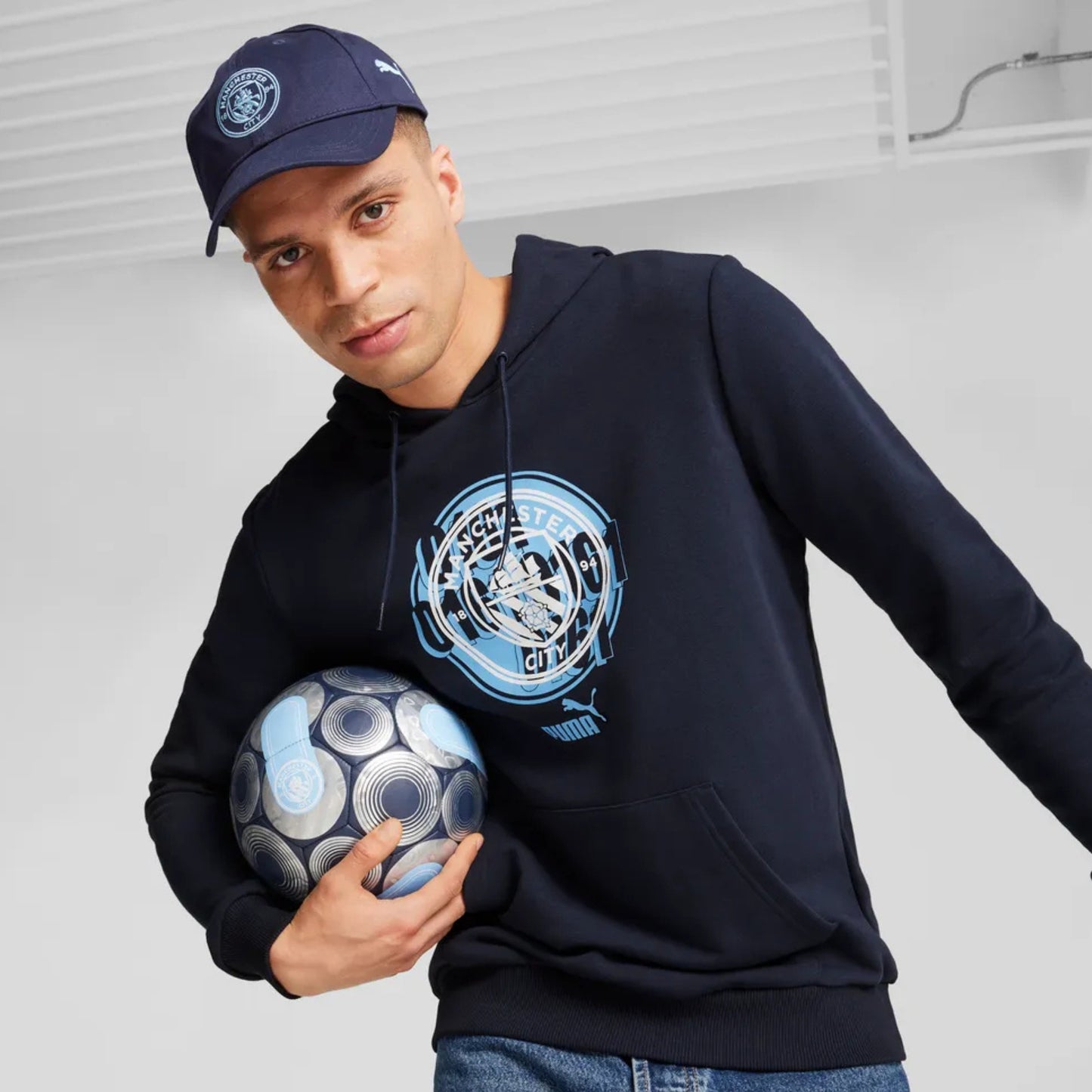 Man wearing a Puma Manchester navy cap holding a soccer ball against a plain background