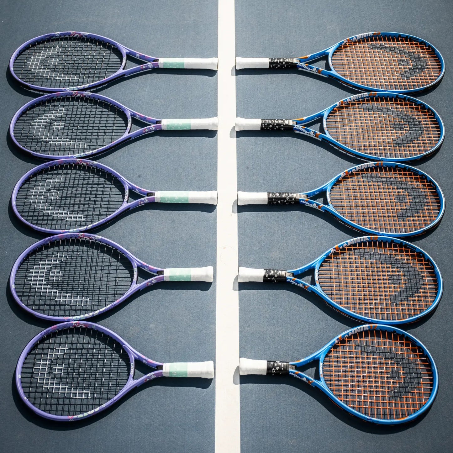 Eight Head tennis rackets arranged in two vertical rows on a blue tennis court, separated by a white center line. The rackets have purple, blue, and black frames with white and green grip tape, and the strings are orange, black, or white.