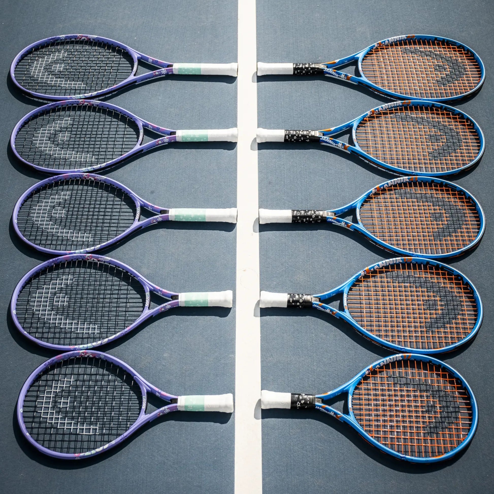 Eight Head tennis rackets arranged in two vertical rows on a blue tennis court, separated by a white center line. The rackets have purple, blue, and black frames with white and green grip tape, and the strings are orange, black, or white.