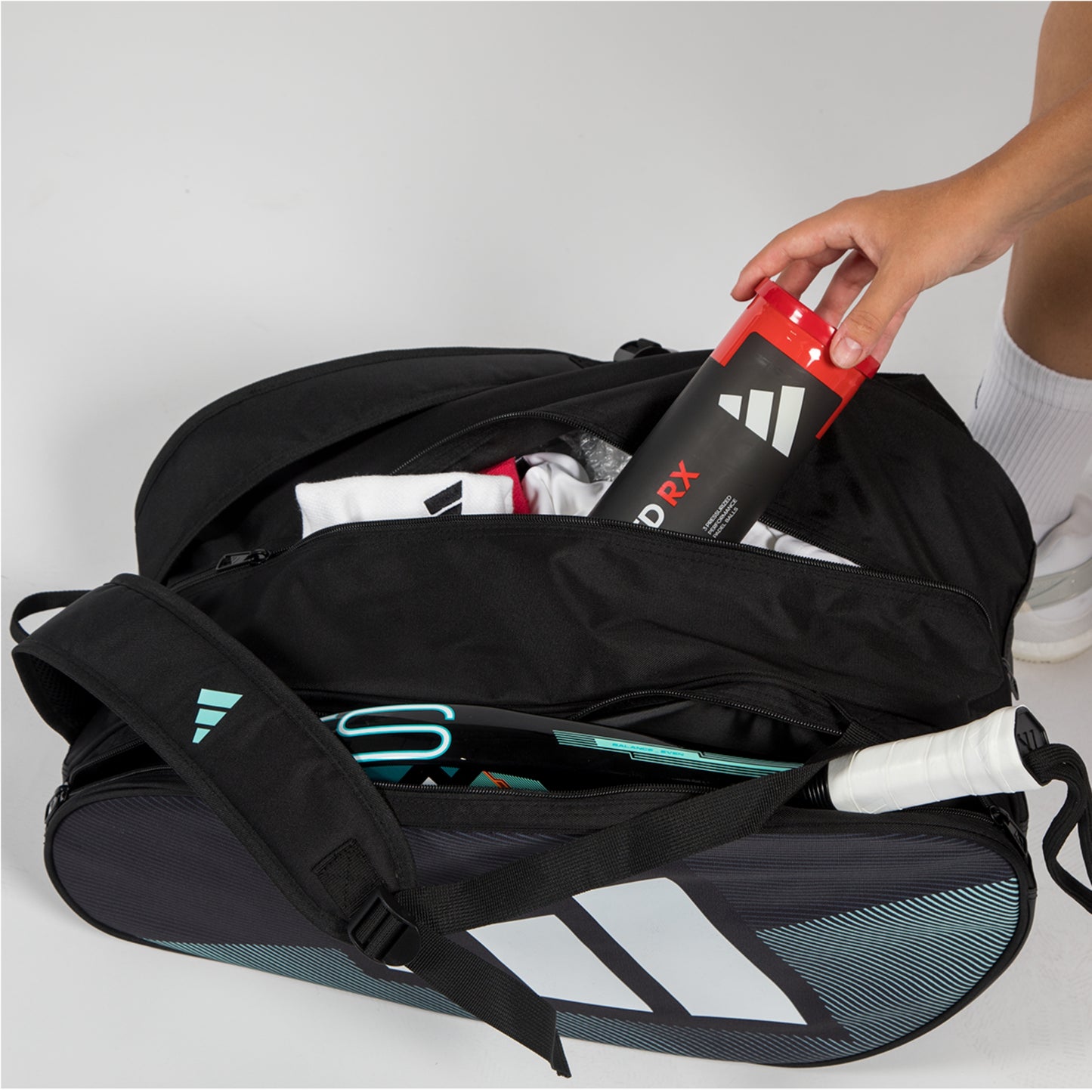 Person opening a black Adidas tennis bag with a red and black shoe inside on a light gray background.