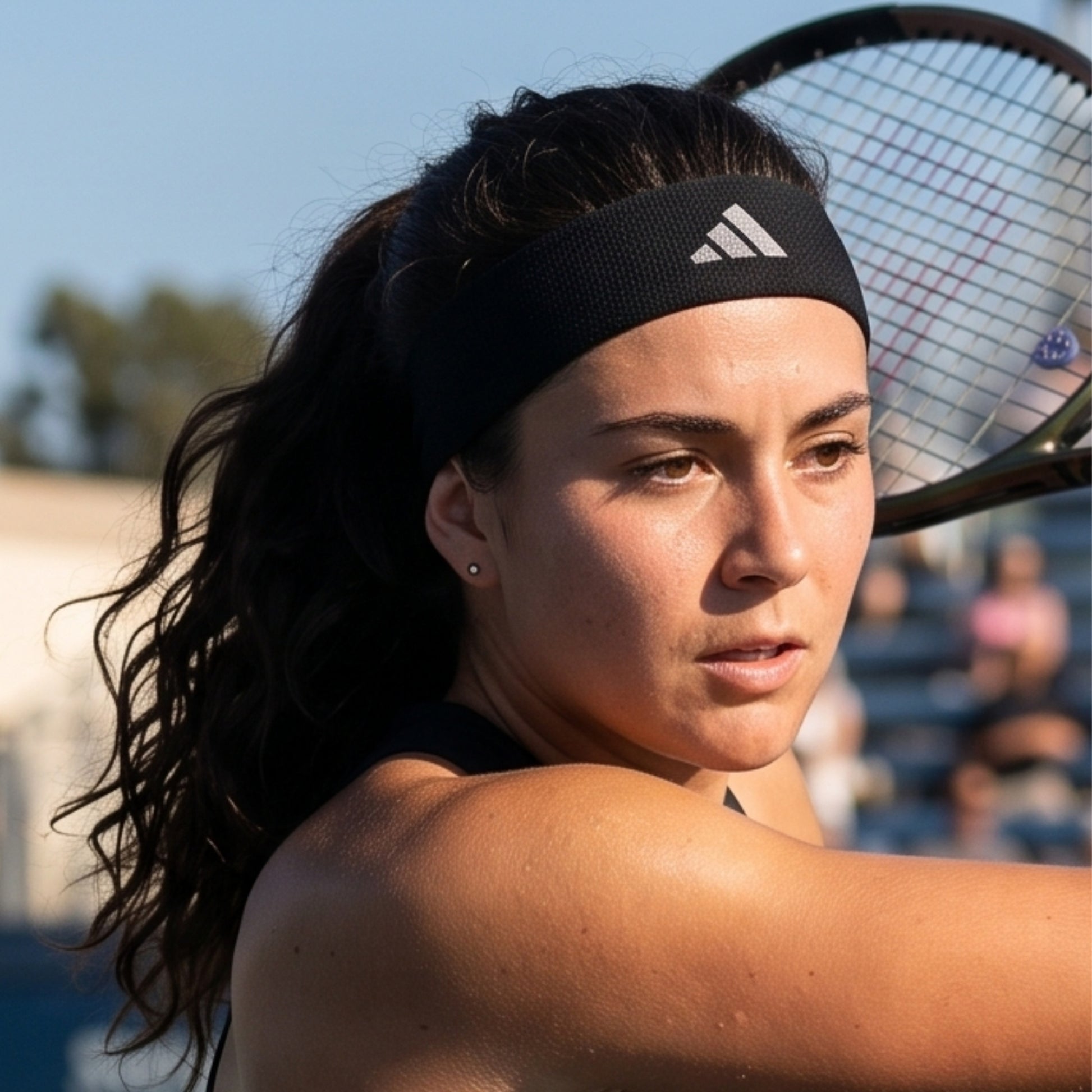 A close-up shot of a female tennis player wearing a black Adidas headband and holding a tennis racket, captured on an outdoor court.