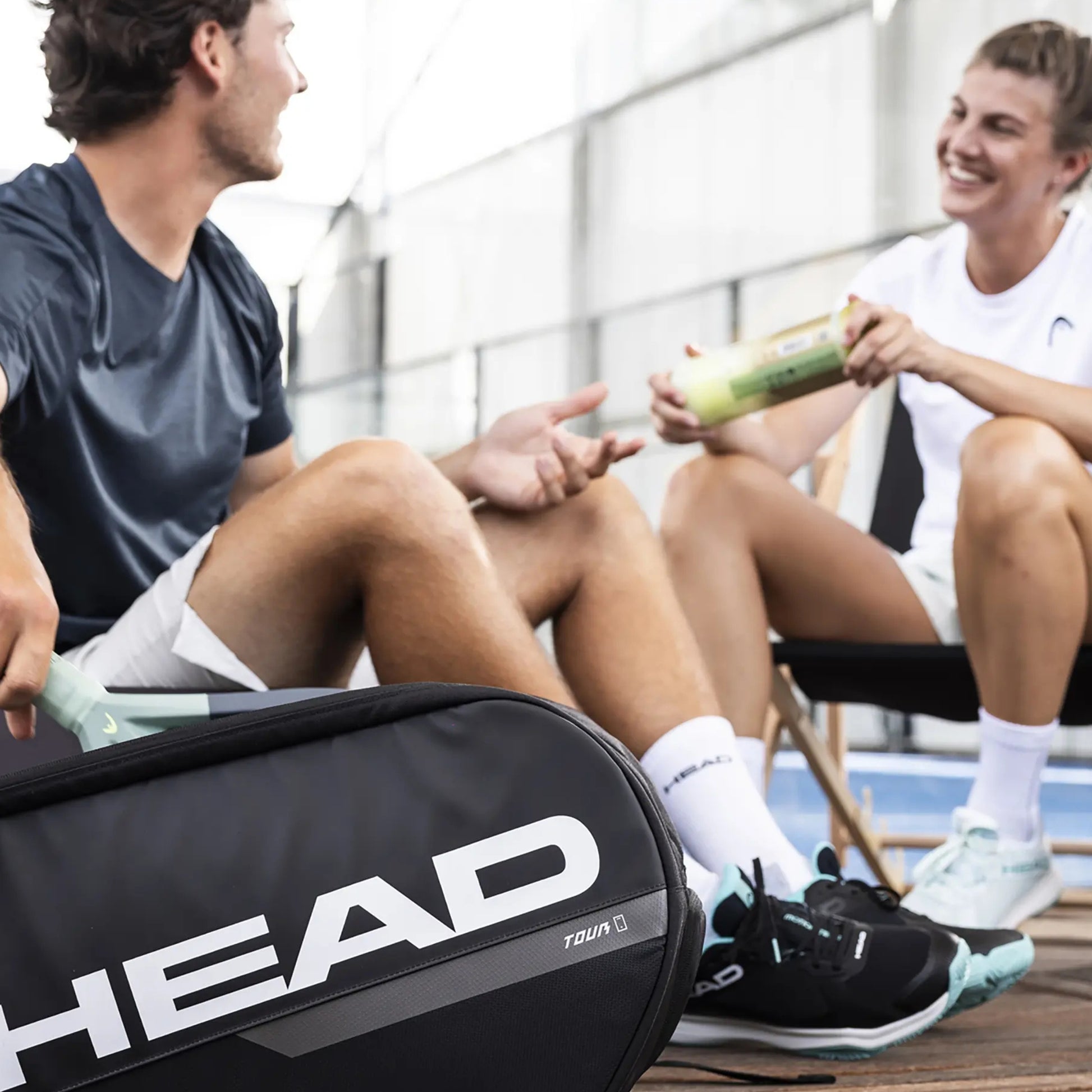 Two people sitting on a bench with a tennis racket case labeled 'HEAD' in the foreground.