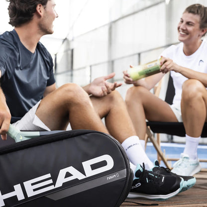Two people sitting on a bench with a tennis racket case labeled 'HEAD' in the foreground.