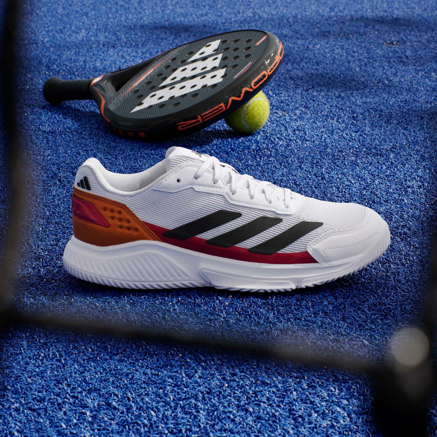 White padel shoe with red and black accents on a blue textured surface, with a paddle and green ball in the background.