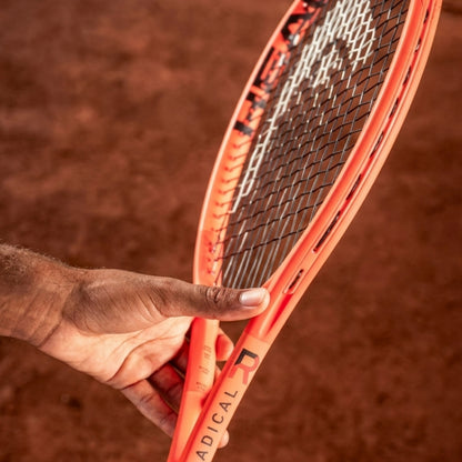 A close-up of a Head tennis racket with a bright orange frame and black-and-white string bed, held by a person's hand against a blurred brown clay court background.