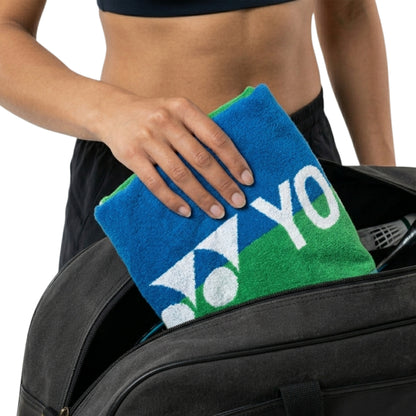 Person holding a blue and green towel with 'YONEX' text, standing next to a suitcase.
