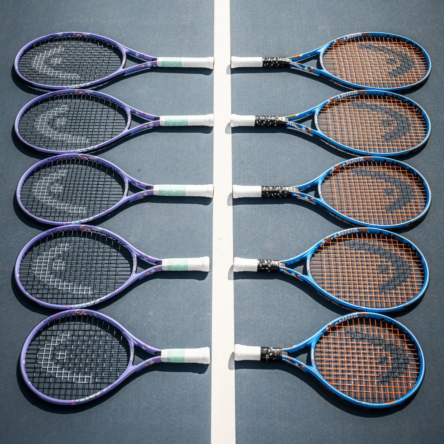 Six Head tennis rackets arranged in two vertical rows on a blue tennis court, with purple, blue, black, and orange racket frames and strings.