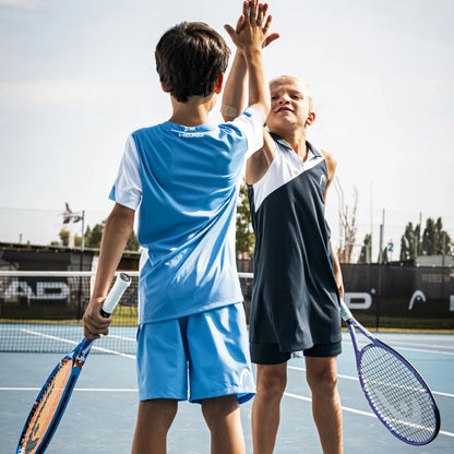 Two children playing tennis on an outdoor court, high-fiving while holding tennis rackets. One child wears a light blue and white tennis set, and the other wears a dark navy and white sleeveless set.