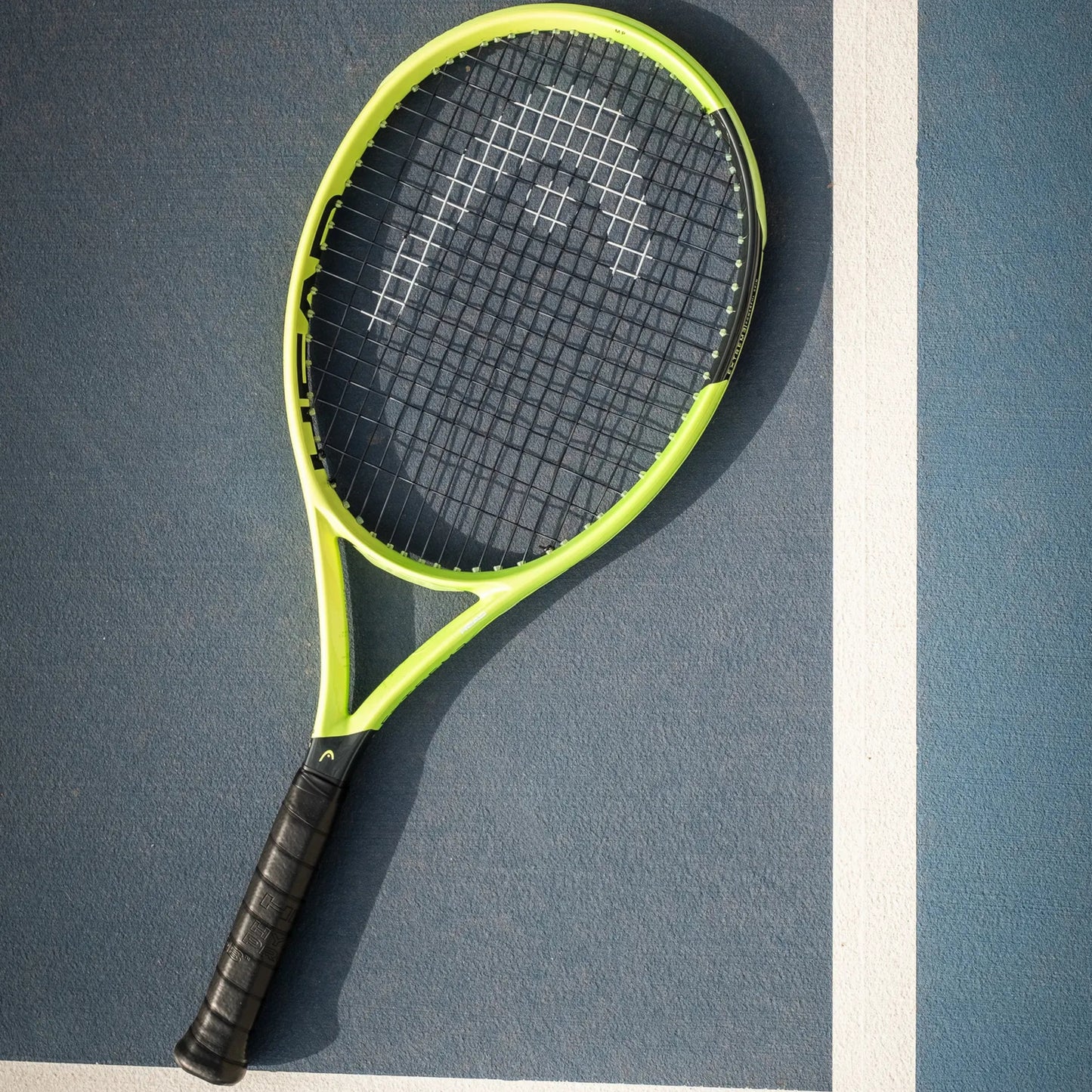 A Head tennis racket with a bright neon yellow-green frame and black grip, resting diagonally on a blue tennis court with a white boundary line.