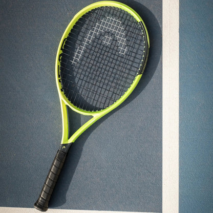 A Head tennis racket with a bright neon yellow-green frame and black grip, resting diagonally on a blue tennis court with a white boundary line.