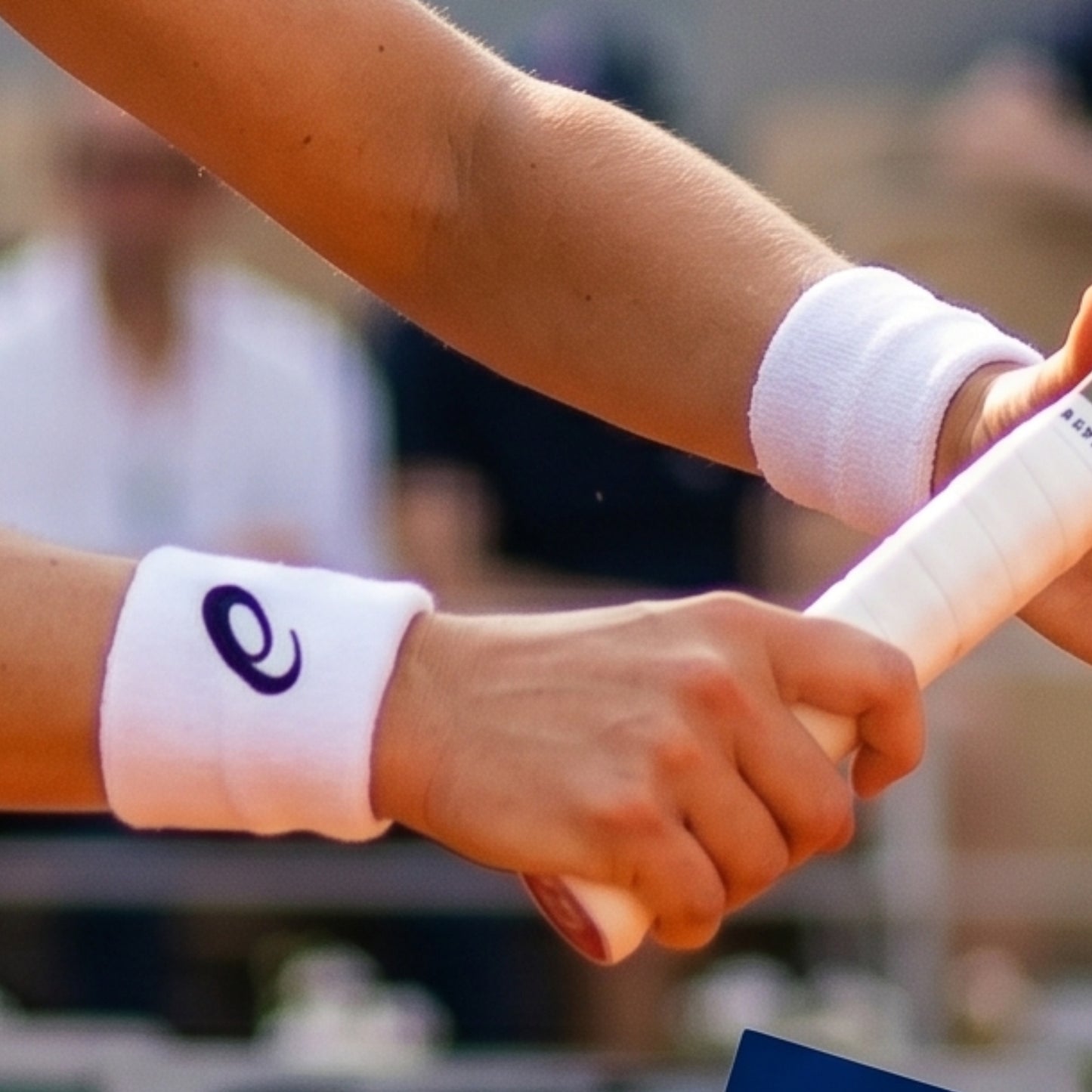 Two hands holding a tennis racket with white wristbands featuring a logo, blurred background