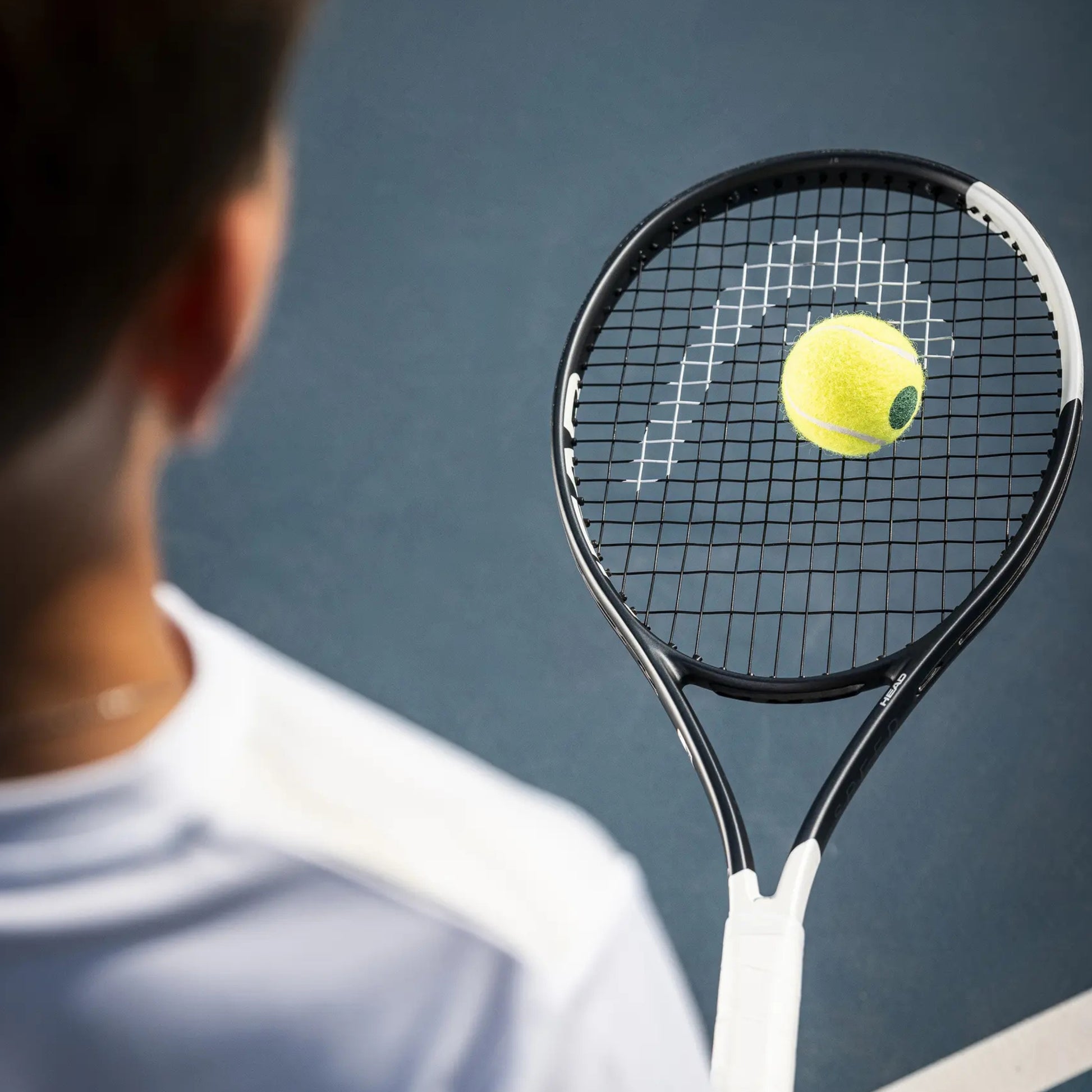 A tennis racket with a yellow tennis ball on its strings, shown from behind a blurred player on a blue tennis court.