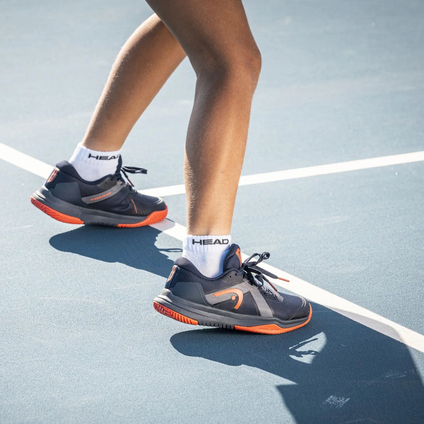 Person wearing black and orange Head padel shoes with white socks on a tennis court.